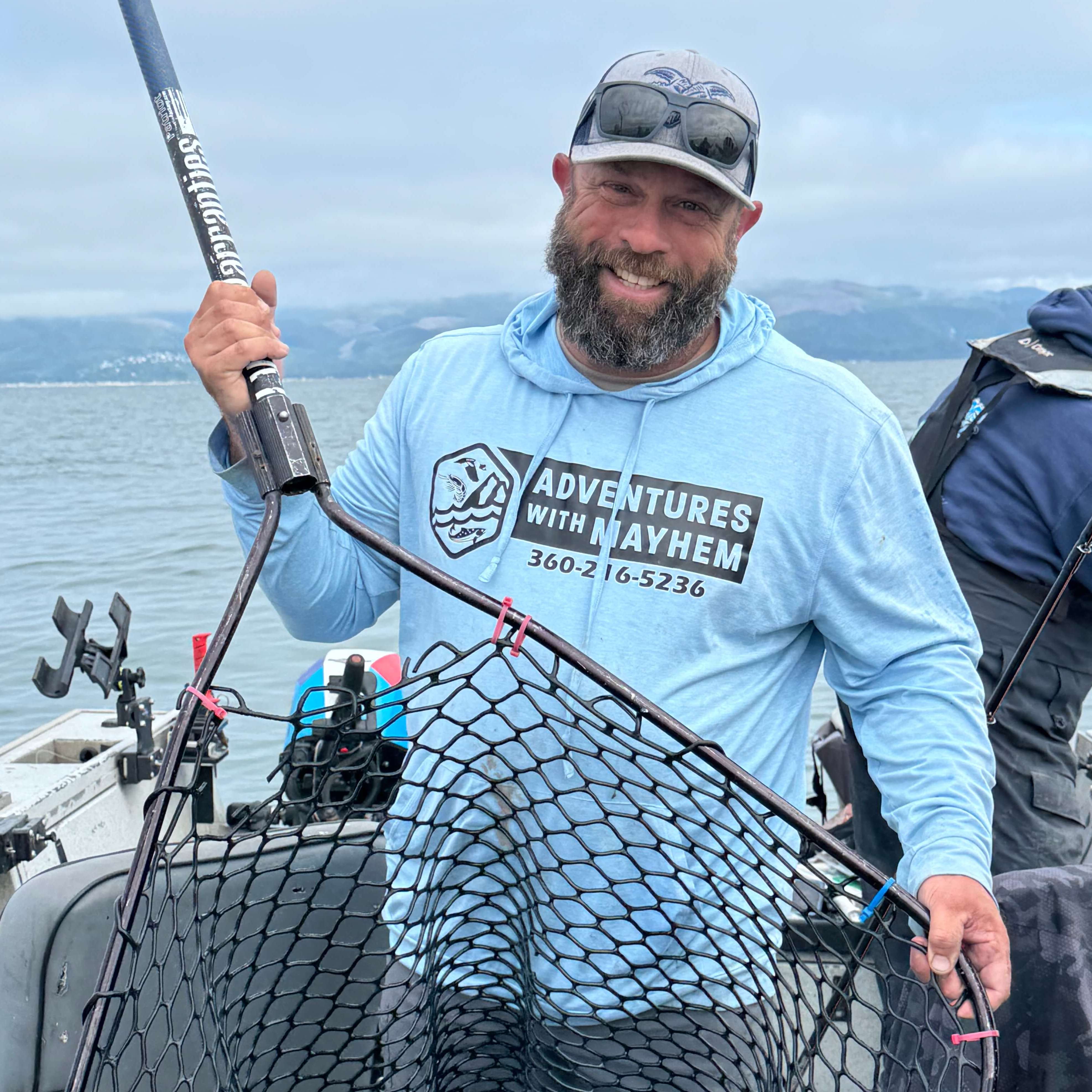 Fishing guide Branden Trager of Adventures with Mayhem Outfitters holding a landing net on a guided salmon trip in Oregon, wearing branded hoodie with coastal mountains in the background.