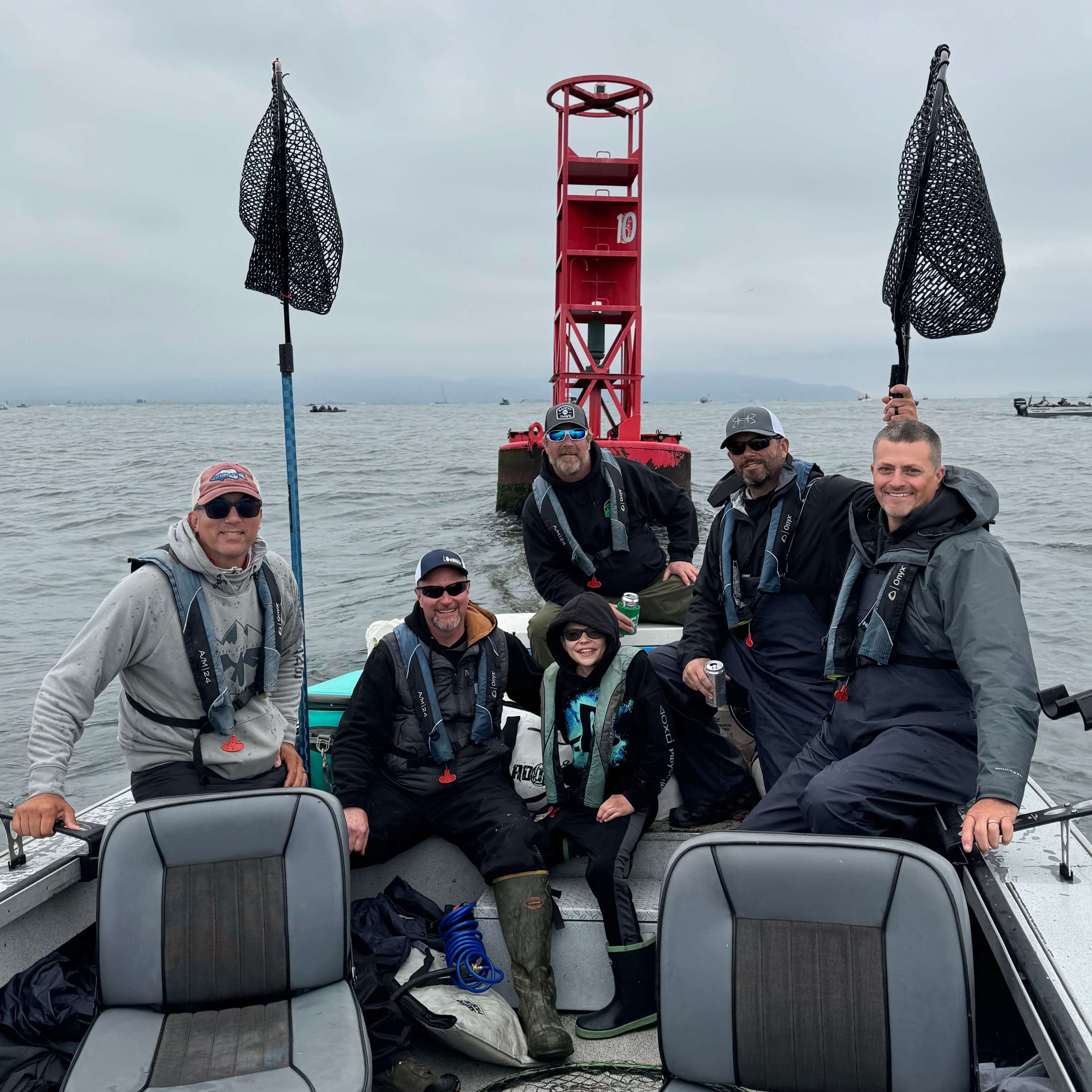 Group of anglers smiling on a guided salmon fishing trip at Buoy 10 near the Columbia River mouth, with red channel marker and landing nets visible—Mayhem Outfitters adventure in Astoria, Oregon.