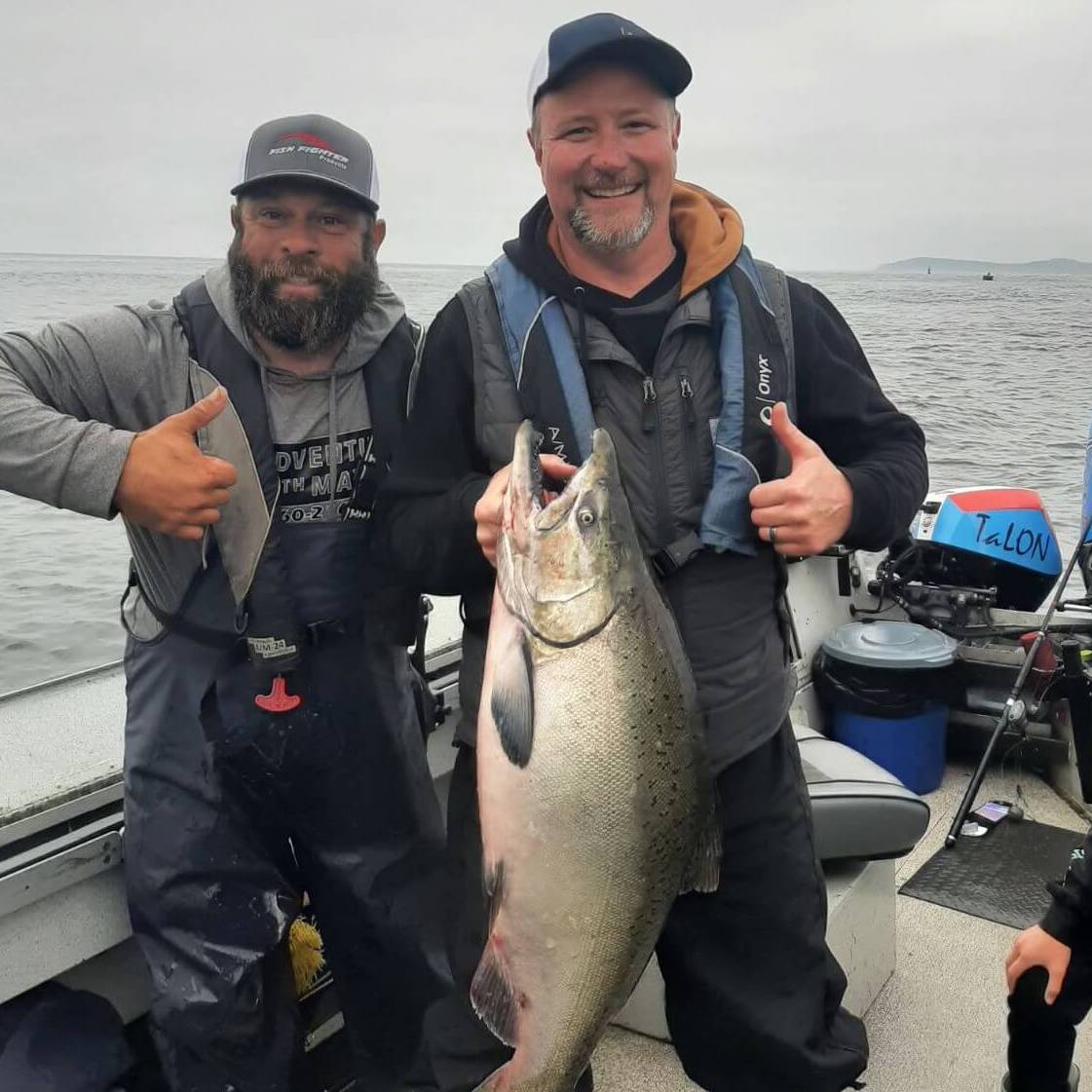 Two anglers smiling on a guided ocean salmon fishing trip in Oregon, holding a massive Chinook salmon aboard a Mayhem Outfitters boat with Pacific waters in the background.