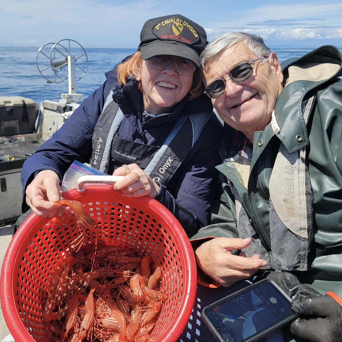 Smiling couple on a guided ocean trip in Oregon holding a red basket full of freshly caught spot prawns—Adventures with Mayhem seafood charter experience.